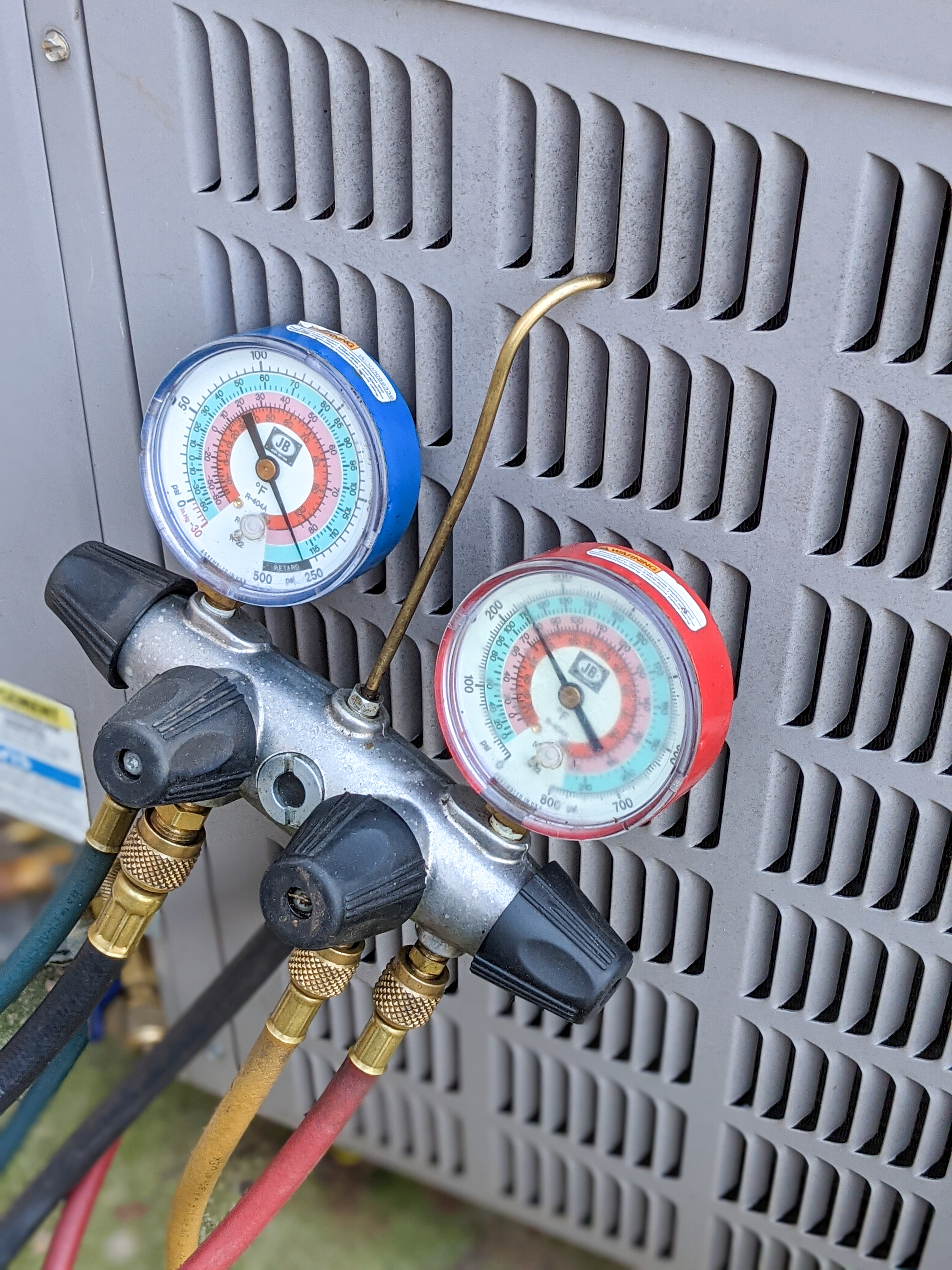 HVAC manifold gauge set connected to an outdoor air conditioning unit, measuring refrigerant pressure for system diagnostics and repair.